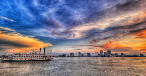 A riverboat on calm waters under a vibrant sunset with colorful clouds and a city skyline in the background.