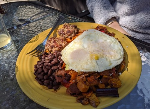 A yellow plate with a fried egg, black beans, sweet potatoes, and sausage patties, set on a glass table.