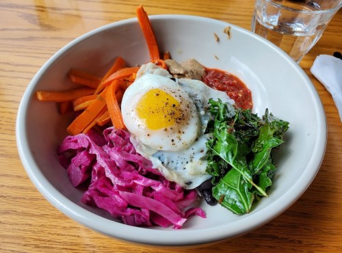 A bowl with sautéed carrots, purple cabbage, greens, meat, and a fried egg on top, served with a side of sauce.