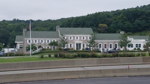 A large stone building with a green roof, surrounded by trees and grass, set against a hilly background.
