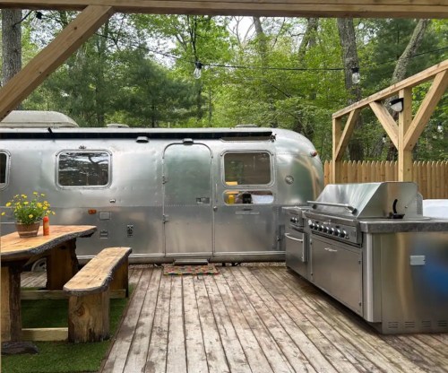 A silver Airstream trailer beside a wooden deck with a grill and picnic table, surrounded by trees.