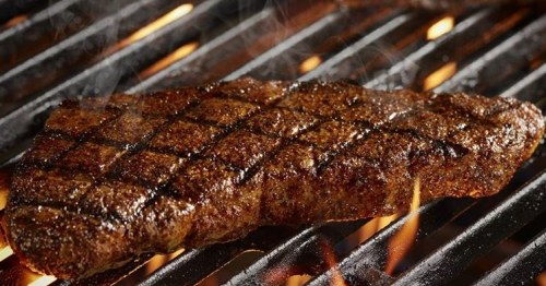 Grilled steak with a seared crust, resting on a hot grill with flames and smoke in the background.