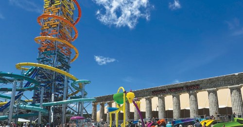 A colorful water slide tower stands tall against a blue sky, with playful water features and umbrellas below.