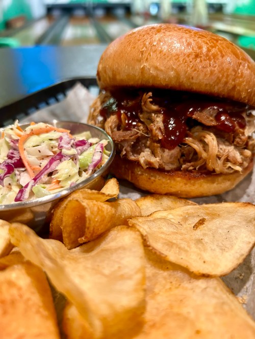 A pulled pork sandwich with BBQ sauce, served with coleslaw and crispy potato chips on the side.