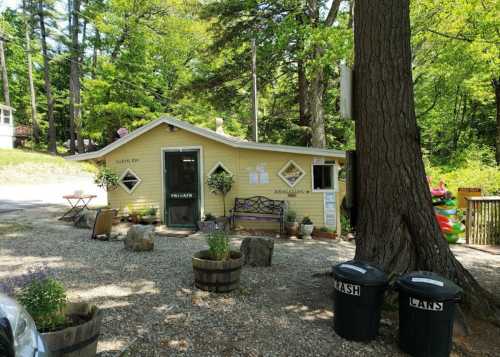 A small yellow cottage surrounded by trees, with a gravel path, benches, and trash cans nearby.