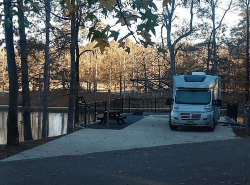 A white RV parked near a serene lake surrounded by trees in autumn. A picnic area is visible nearby.