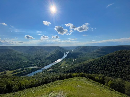 A panoramic view of a river winding through green mountains under a bright blue sky with scattered clouds.