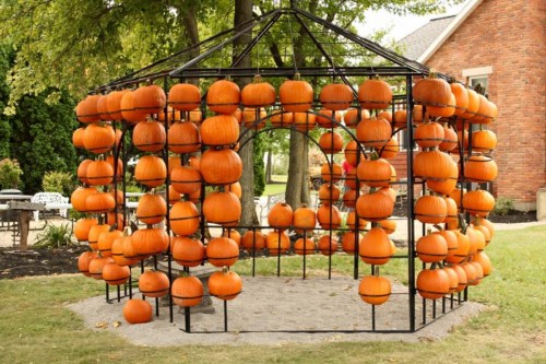 A decorative structure filled with numerous orange pumpkins, set in a grassy area near a brick building.