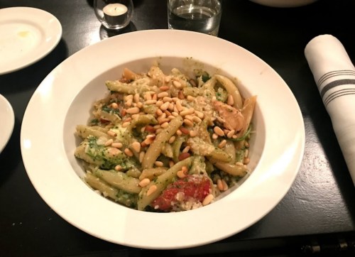 A bowl of pasta with pesto, artichokes, sun-dried tomatoes, and pine nuts, served on a black table.