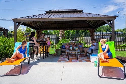 A group of people relaxes under a gazebo, enjoying drinks and snacks on a sunny day by a poolside.