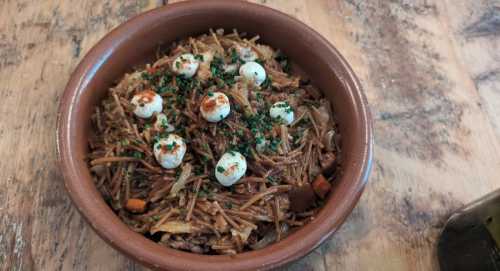 A bowl of seasoned noodles topped with small cheese balls and herbs, served on a wooden table.