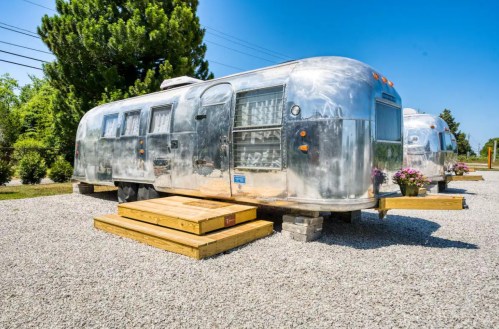 Two vintage Airstream trailers parked on a gravel lot, surrounded by greenery and wooden platforms.