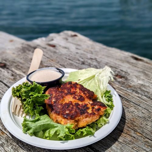A grilled fish fillet on a bed of lettuce, served with a small cup of sauce, on a rustic wooden table by the water.