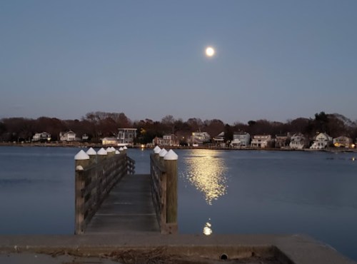 A serene waterfront scene at dusk, featuring a wooden pier and a full moon reflecting on the calm water.