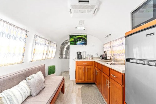 Interior of a modern camper with a sofa, kitchen area, and a TV, featuring bright curtains and wooden cabinetry.