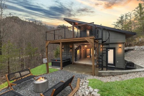 Modern cabin with a fire pit, surrounded by trees and mountains, featuring a deck and large windows at sunset.