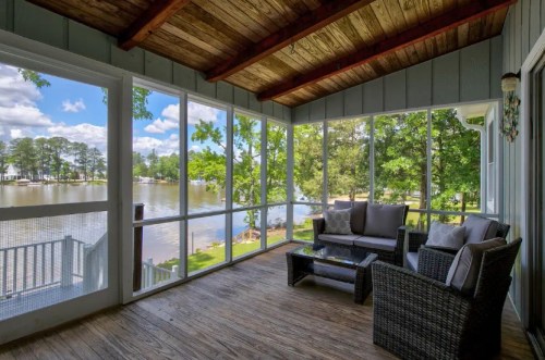 A cozy screened porch with a view of a lake, featuring a seating area and natural light streaming in.
