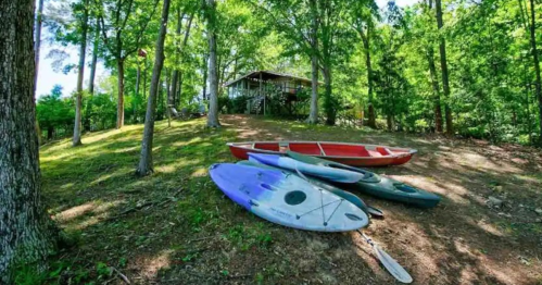 A collection of kayaks on a grassy area, with a cabin nestled among trees in the background.