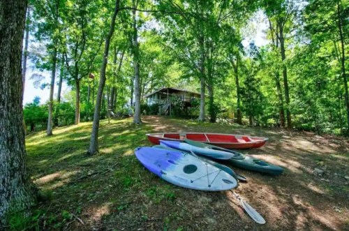 A serene scene of kayaks and canoes on a grassy slope, with a cabin nestled among lush green trees in the background.