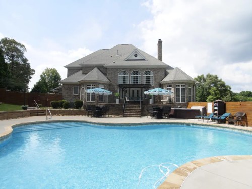 A large house with a stone exterior overlooks a blue swimming pool, surrounded by patio furniture and umbrellas.