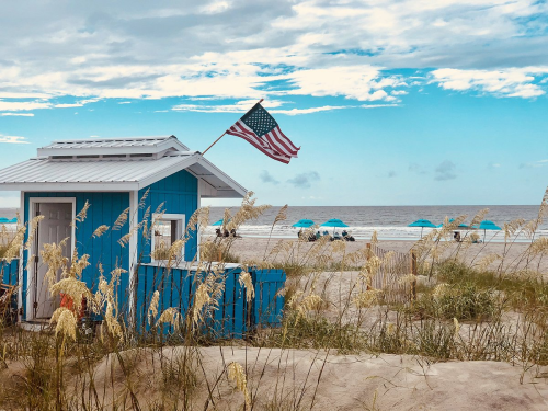 A blue beach hut with an American flag, surrounded by tall grass and umbrellas on a sandy beach under a cloudy sky.