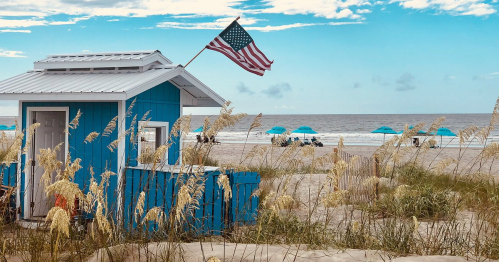 A blue beach hut with an American flag, surrounded by tall grass and umbrellas on a sandy beach under a blue sky.