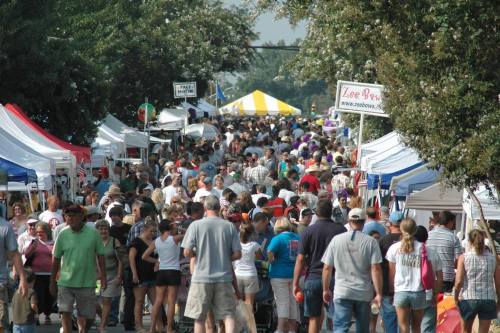 A bustling outdoor market with crowds of people walking between colorful vendor tents under a clear sky.