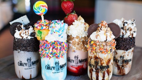 Five colorful, extravagant milkshakes topped with whipped cream, sprinkles, candy, and fruit, displayed on a table.