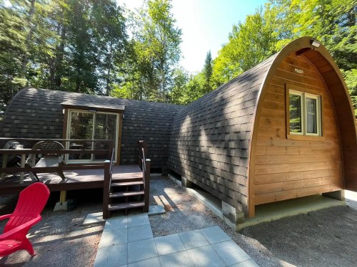 A unique, curved wooden cabin surrounded by trees, featuring a deck and red chairs in a serene outdoor setting.