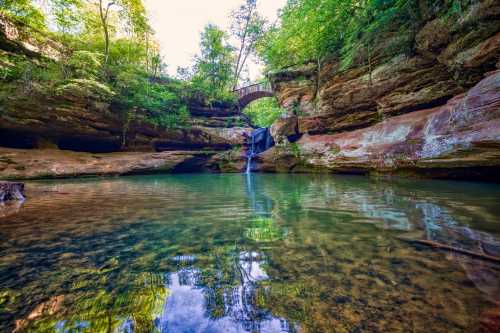 A serene waterfall cascades into a clear pool, surrounded by lush greenery and a wooden bridge in the background.