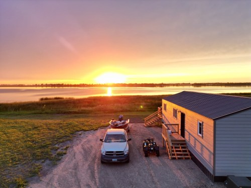 A serene sunset over a lake, with a parked truck, a kayak, and an ATV near a cozy cabin.