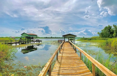 A wooden dock leads to green structures over a calm, reflective lake surrounded by lush vegetation and blue skies.