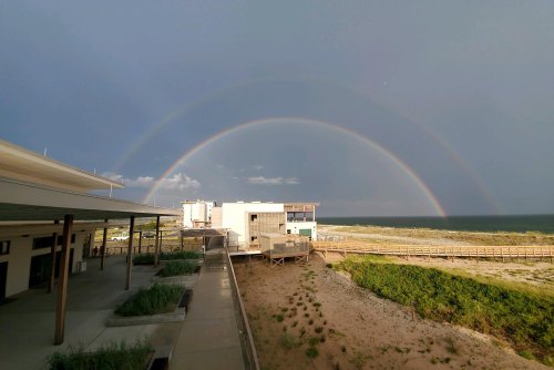 A double rainbow arcs over a beachside building, with sandy dunes and ocean in the background under a cloudy sky.