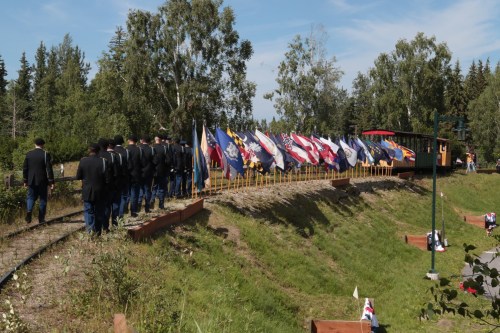 A group of people in uniform walks along a path lined with various flags, surrounded by trees and greenery.