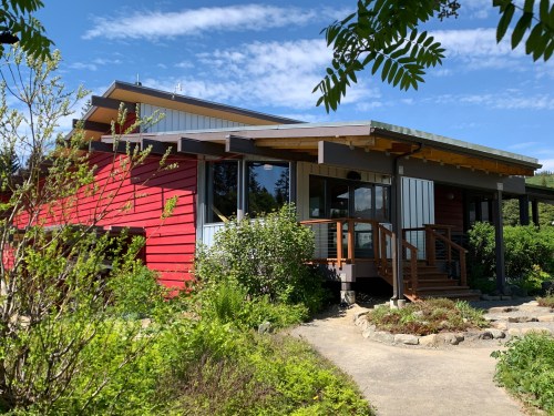 A modern house with red and gray siding, surrounded by greenery and a clear blue sky.
