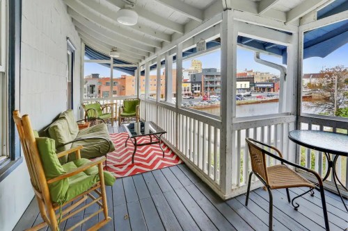 A cozy porch with rocking chairs and a small table, overlooking a waterfront and cityscape.