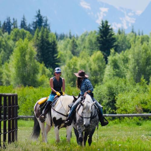 Two women on horseback in a lush green landscape, surrounded by trees and mountains in the background.