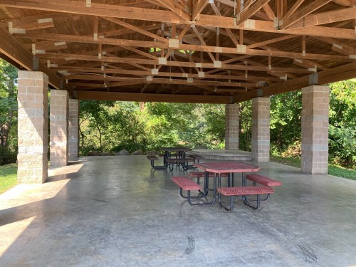 A spacious outdoor pavilion with a wooden roof, featuring picnic tables and surrounded by greenery.