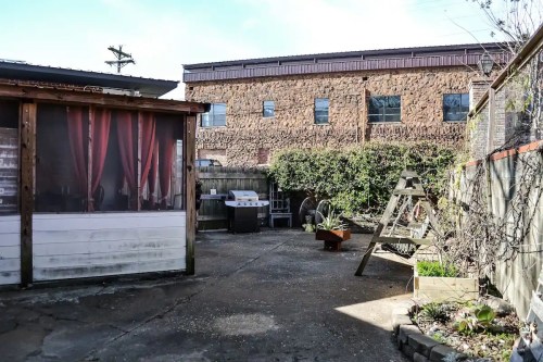 A backyard with a patio area, grill, and greenery, surrounded by a stone building and wooden structures.