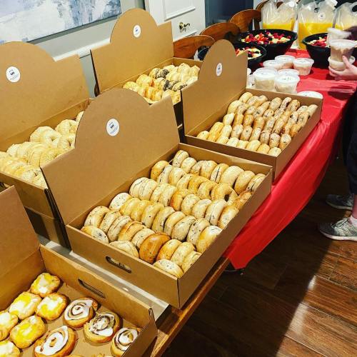 A table with several boxes of assorted bagels and pastries, alongside bowls of fruit and other snacks.