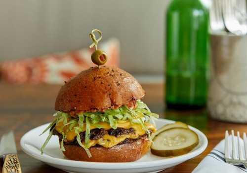 A juicy cheeseburger topped with lettuce, served on a plate with pickles and a green bottle in the background.