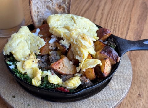A cast iron skillet filled with scrambled eggs, potatoes, sausage, and greens, served with a slice of bread.