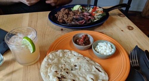 A plate of tortillas with salsa and cheese, alongside a sizzling dish of meat and vegetables, and a drink with lime.