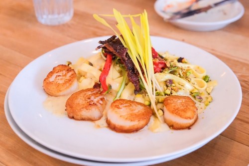 A plate of seared scallops on a bed of pasta with vegetables and garnished with herbs.