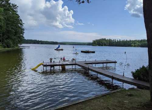 A serene lake scene with a wooden dock, a slide, and people enjoying the water under a partly cloudy sky.