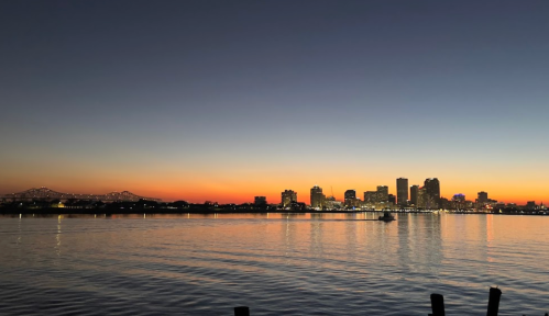 City skyline at sunset, reflecting on calm water with a gradient of orange and blue in the sky.