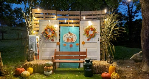 A cozy fall-themed display with a blue door, wreaths, pumpkins, and a bench, set in a park at dusk.