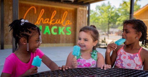Three young girls enjoy colorful popsicles while sitting at a table, with a vibrant "Chakula Snacks" sign in the background.