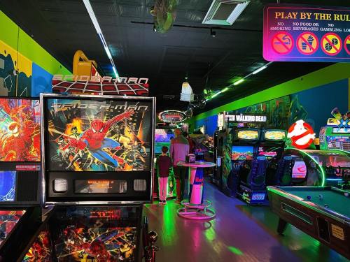 A vibrant arcade filled with various games, including a Spider-Man pinball machine and colorful neon lights.