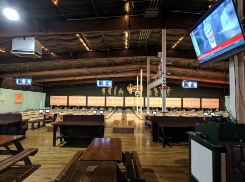 Interior of a bowling alley with lanes, seating, and multiple TVs showing news. The space is well-lit and spacious.